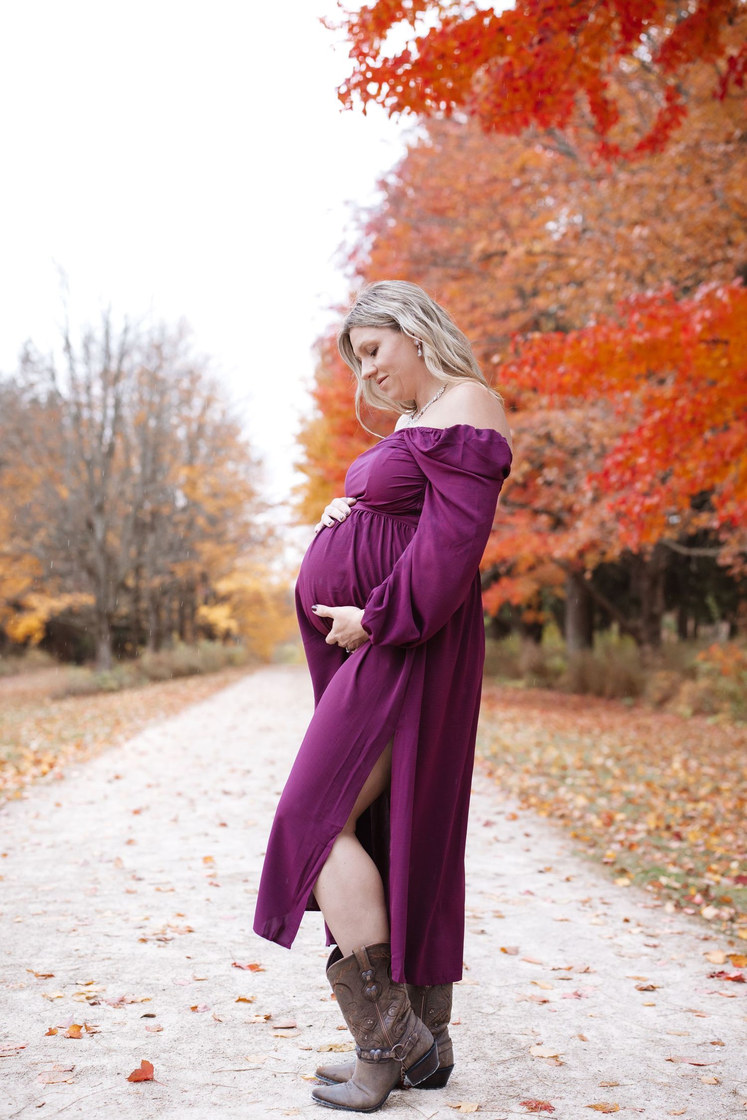 Expectant mother in purple maternity dress and cowboy boots posing on vibrant autumn foliage path.
