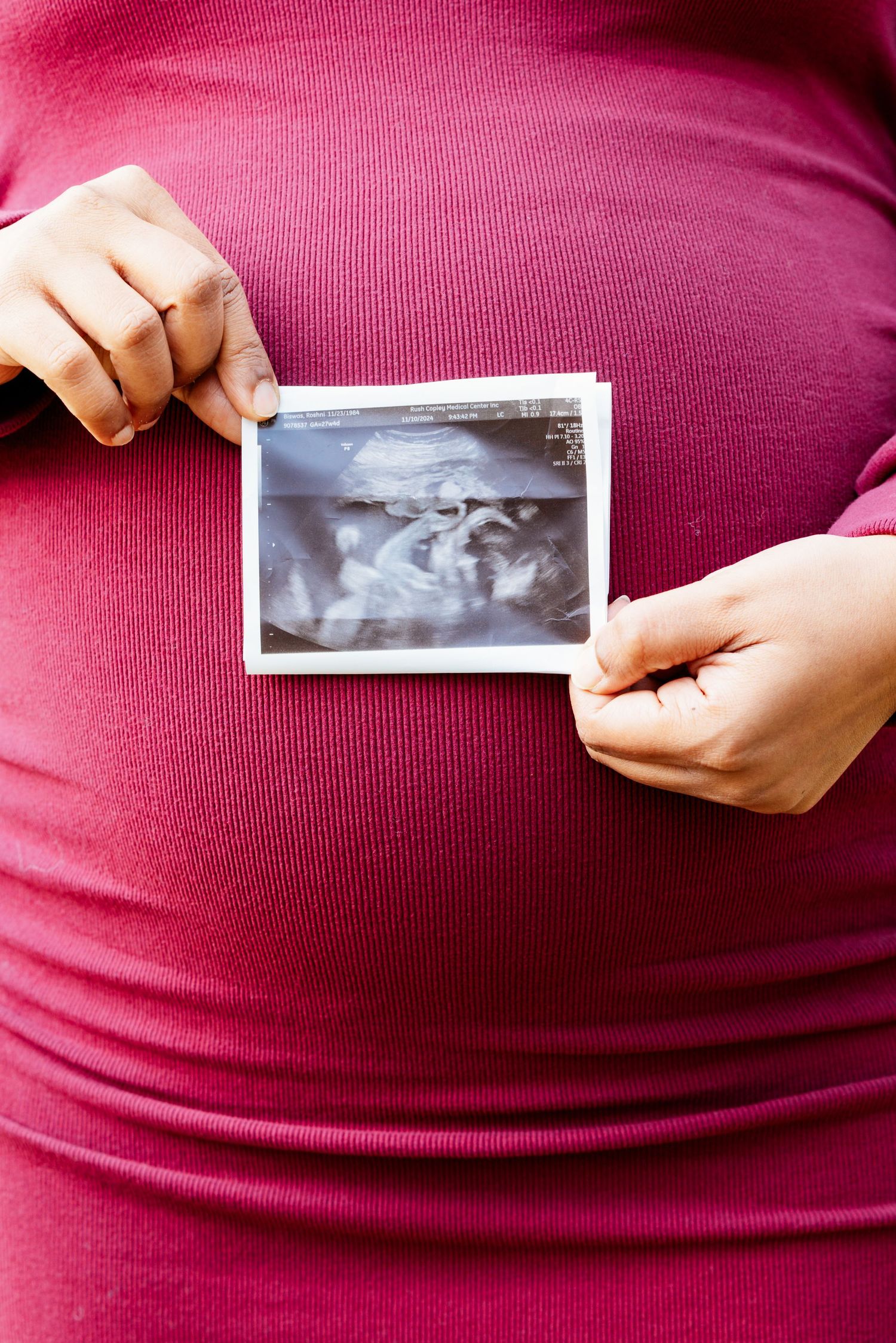Pregnant woman in pink dress holding ultrasound image against her visible baby bump outdoors.