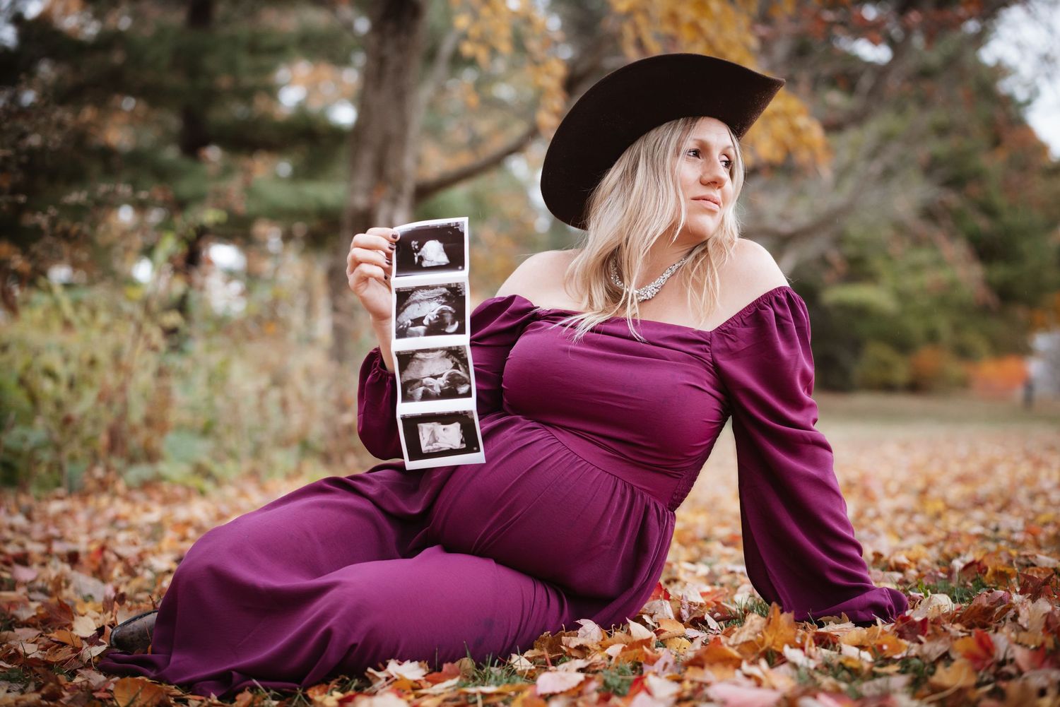 Pregnant woman in purple dress and cowboy hat sitting in autumn leaves holding ultrasound photos outdoors.