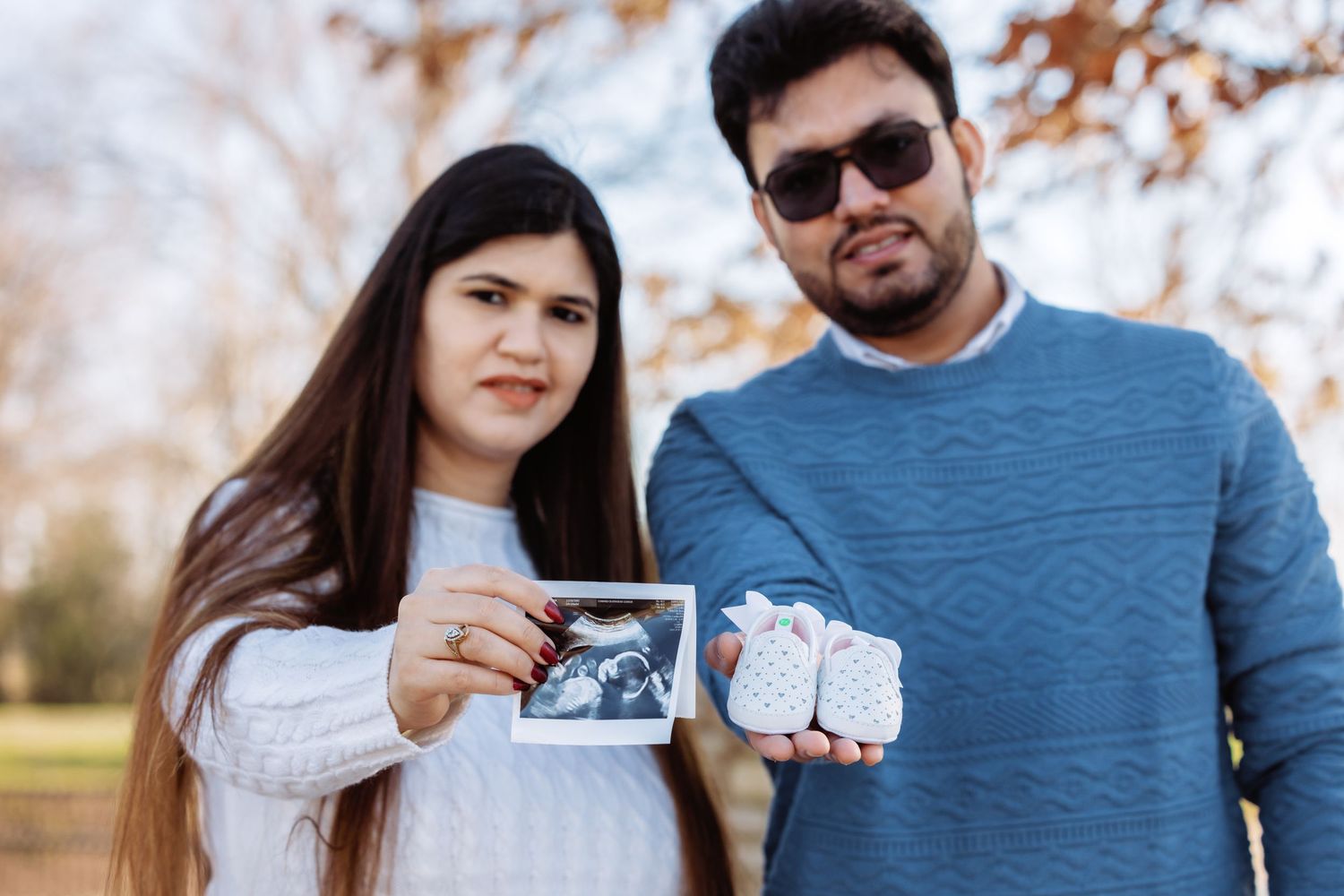 Couple holding ultrasound photo and tiny baby booties outdoors during autumn maternity photoshoot.