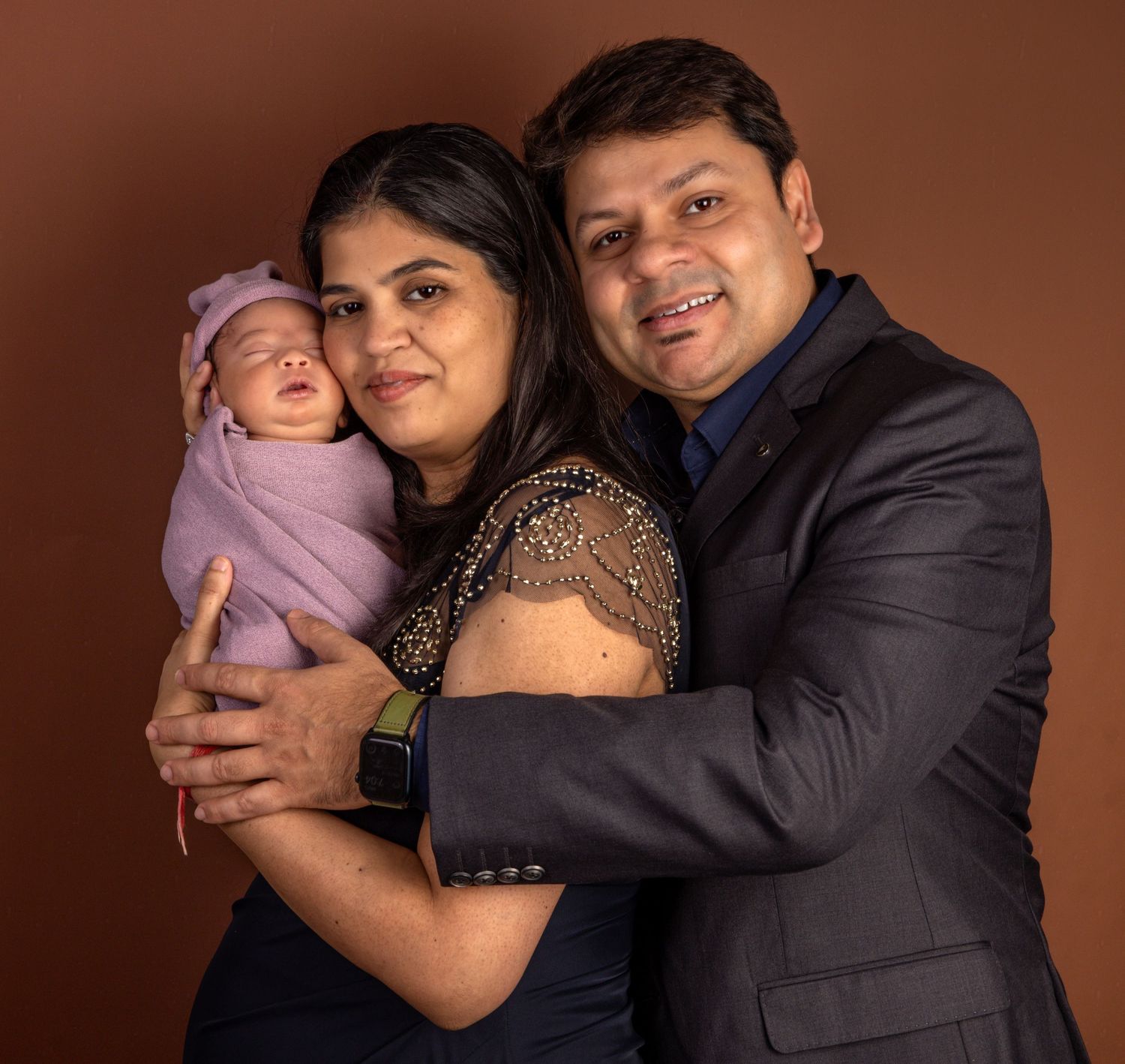 Smiling Indian couple holding swaddled newborn baby in purple wrap against brown background.