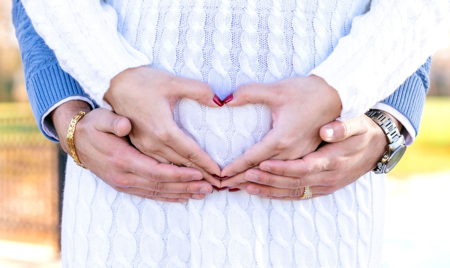 Couple forming heart shape with hands on pregnant belly wearing white knit dress and blue sweater.