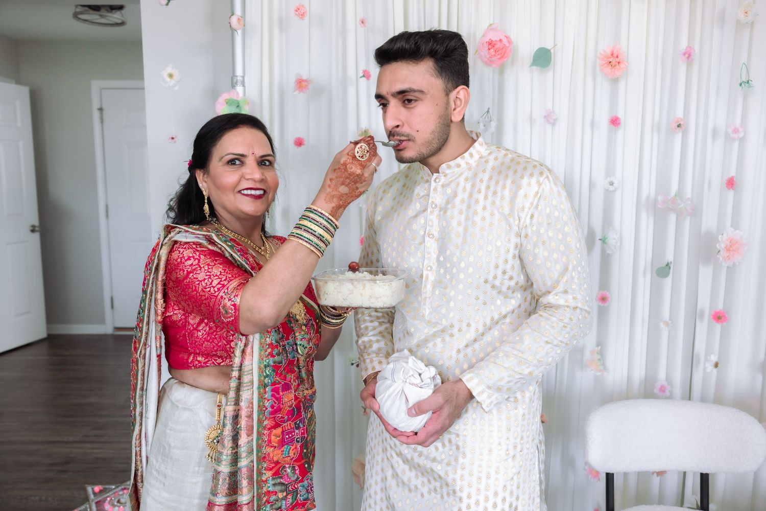 Woman in red saree feeds cake to man in white sherwani at Indian wedding celebration with floral backdrop.