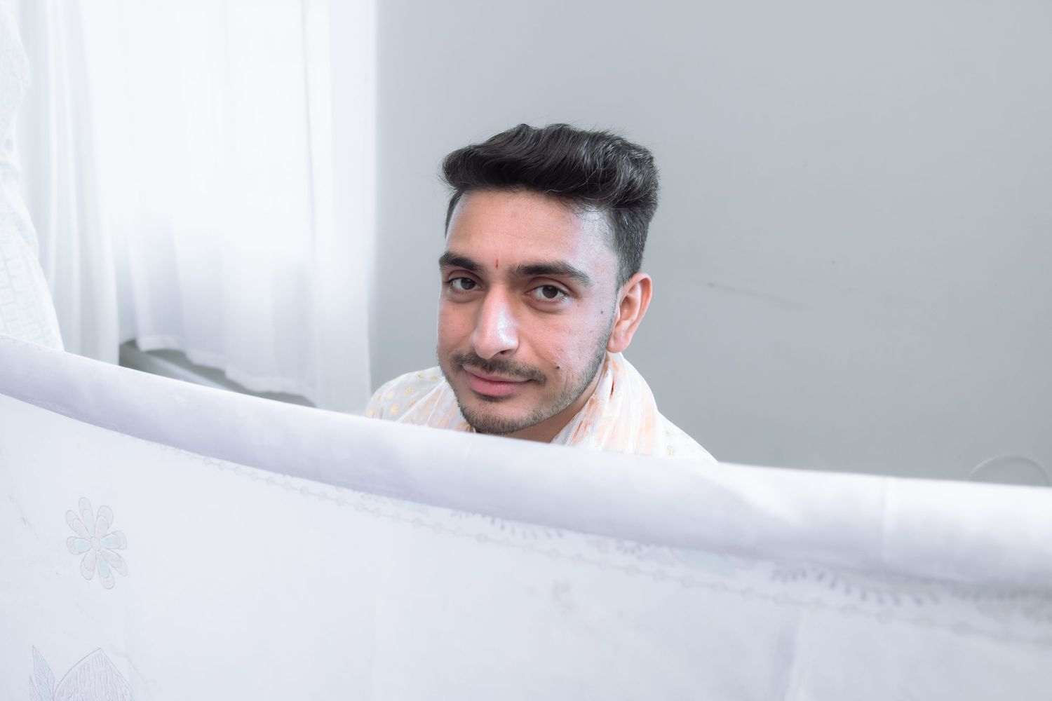 Young South Asian man in white traditional attire smiles warmly at camera during wedding celebration.
