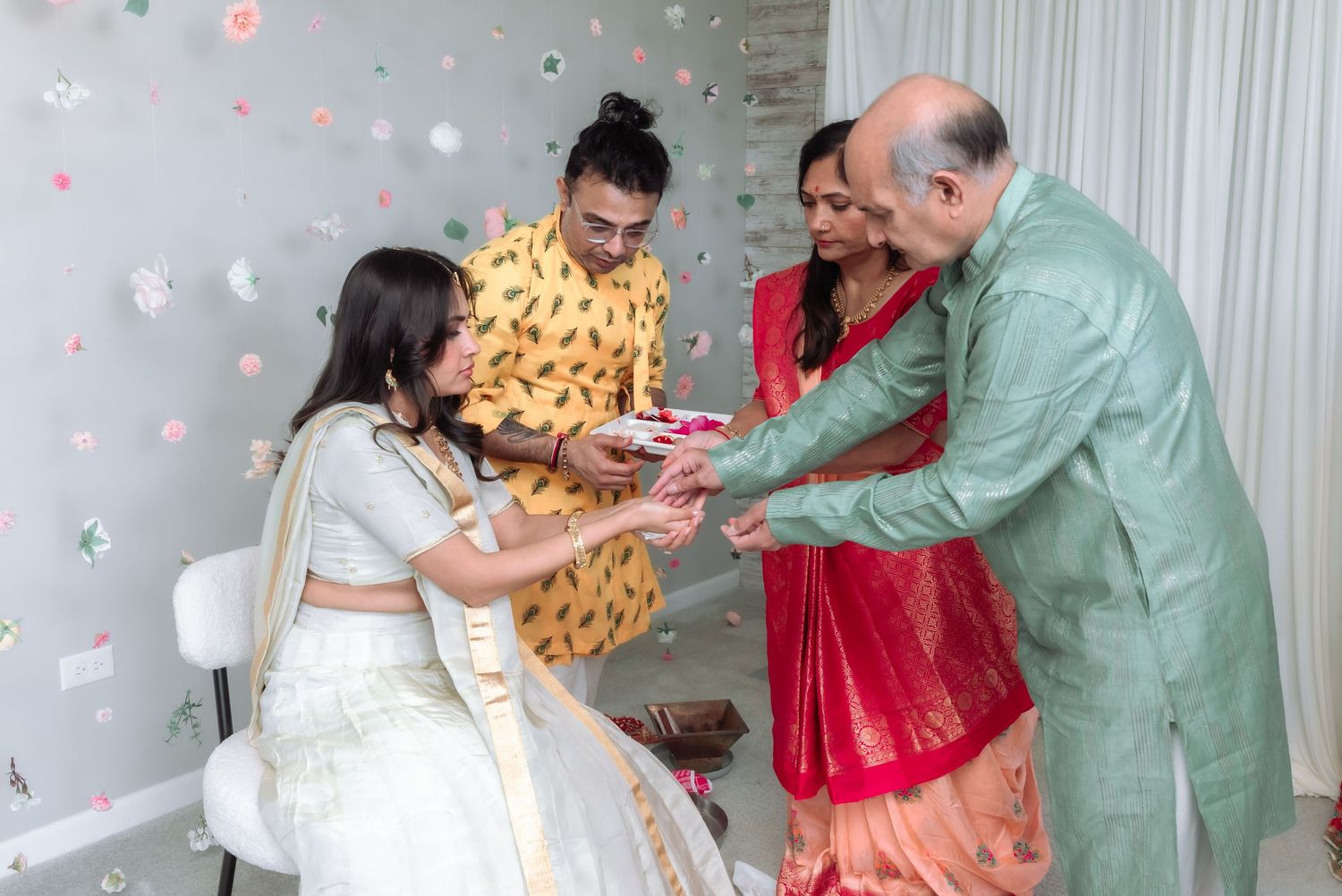A bride in white lehenga receives gifts from family members dressed in colorful Indian traditional wear.
