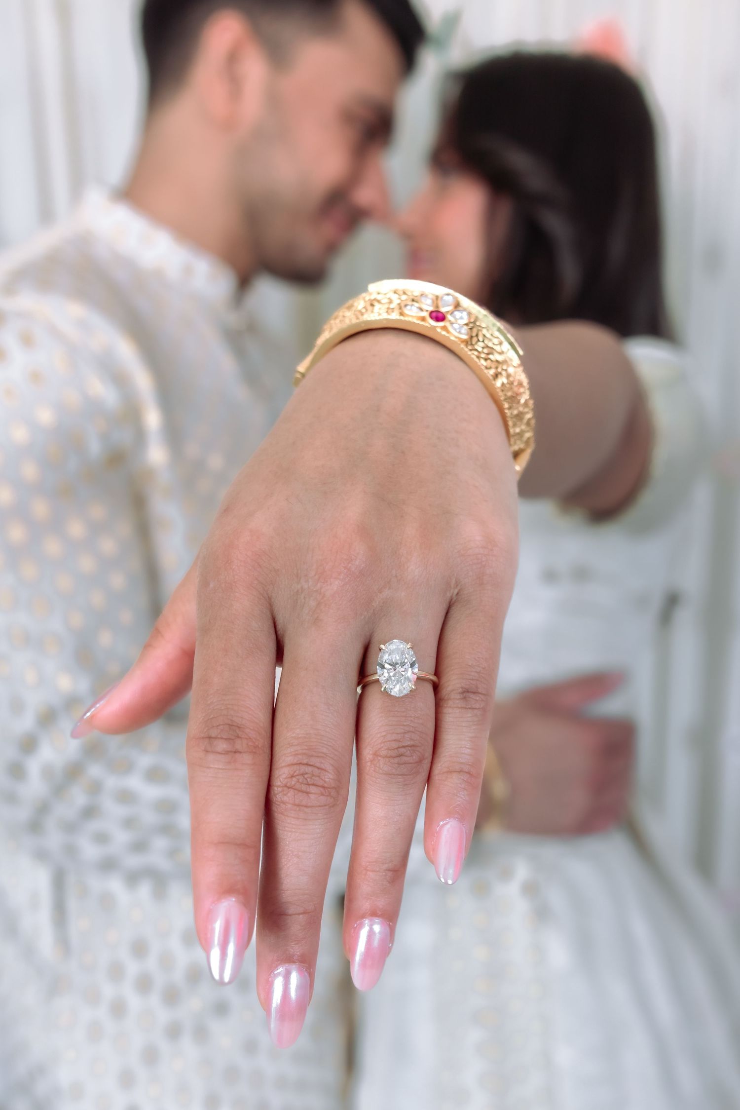 Close-up of diamond engagement ring on bride's hand with couple in white Indian attire in background.