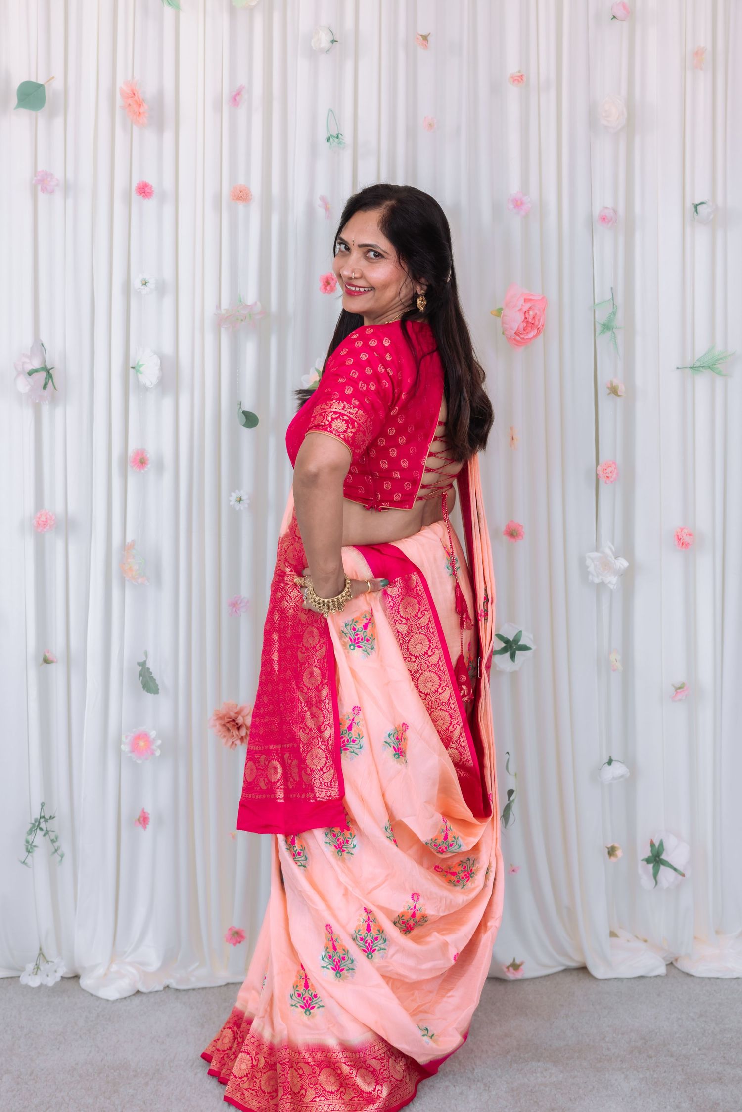 Woman in pink and peach saree posing gracefully before white floral curtain backdrop.