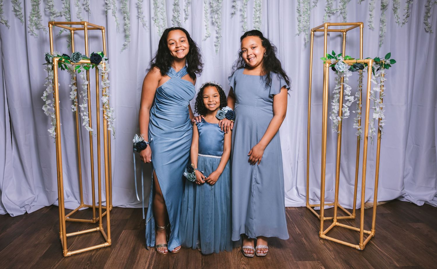 Three girls in dusty blue dresses posing in front of white draped backdrop with gold stands.