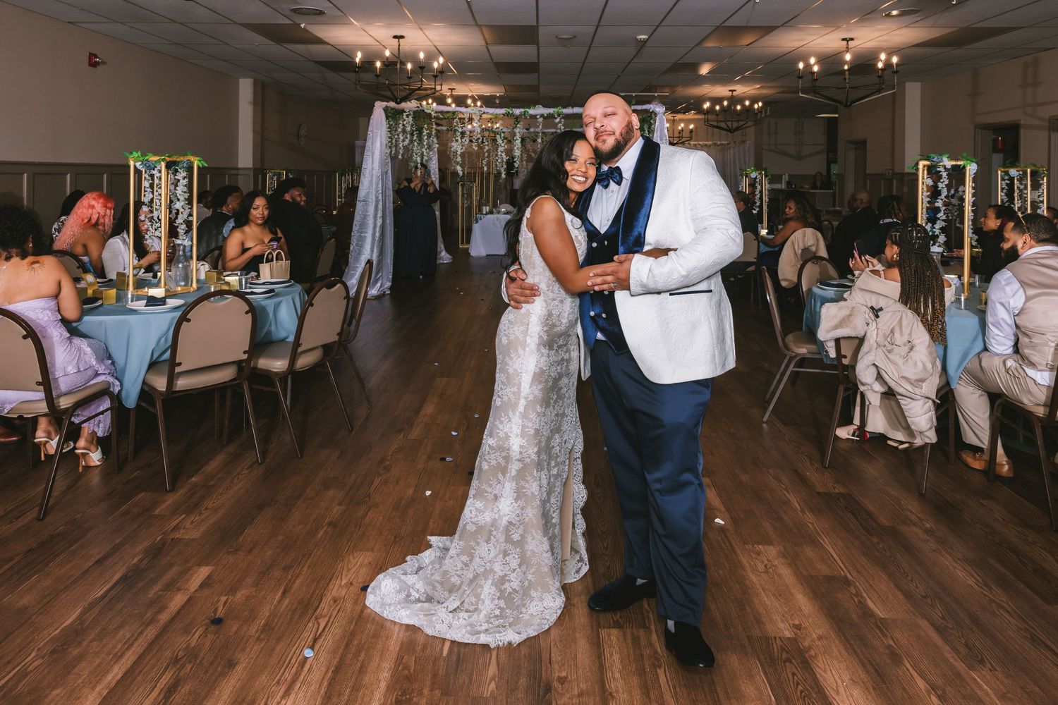 Newlyweds share first dance in elegant wedding reception hall decorated with string lights and floral arrangements.