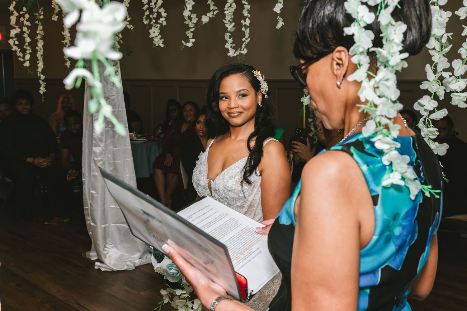 Smiling bride in lace gown with floral hair accessory listens to officiant during indoor wedding ceremony.