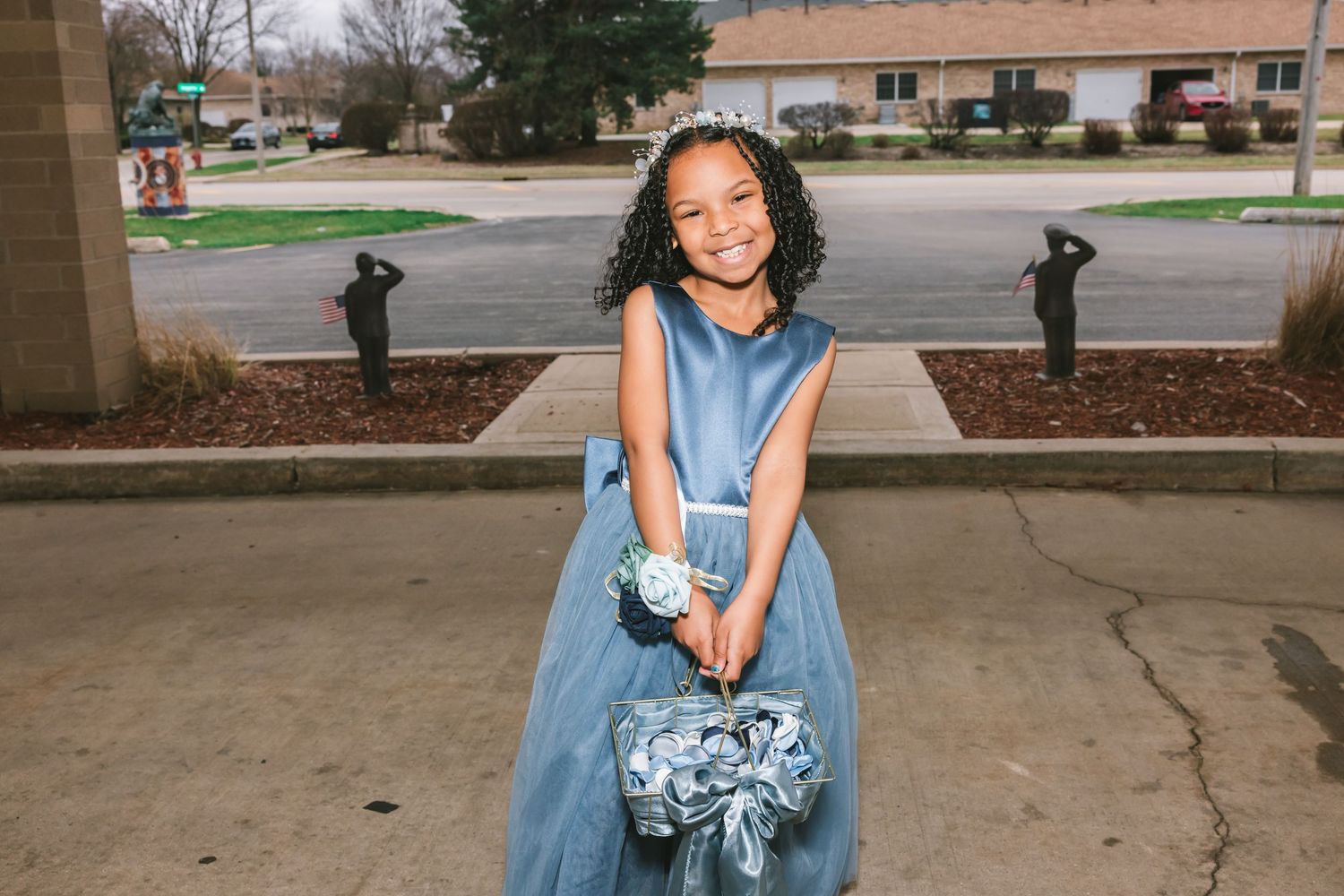 Young flower girl in blue dress holding floral basket smiling outdoors at wedding venue.