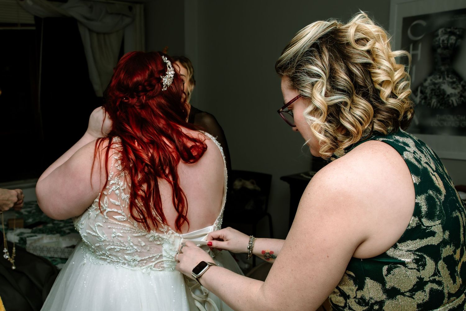 Redhead bride in lace wedding dress being helped by blonde bridesmaid in green dress.