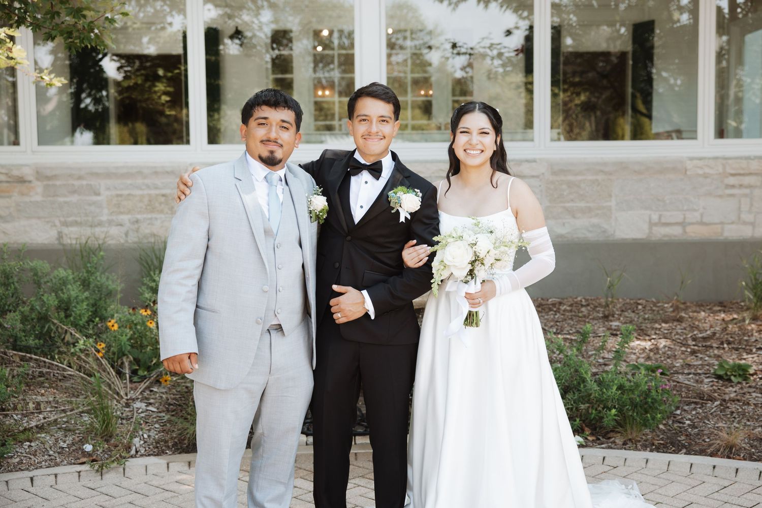 Bride and groom posing with smiling guest outdoors near stone building on their wedding day.