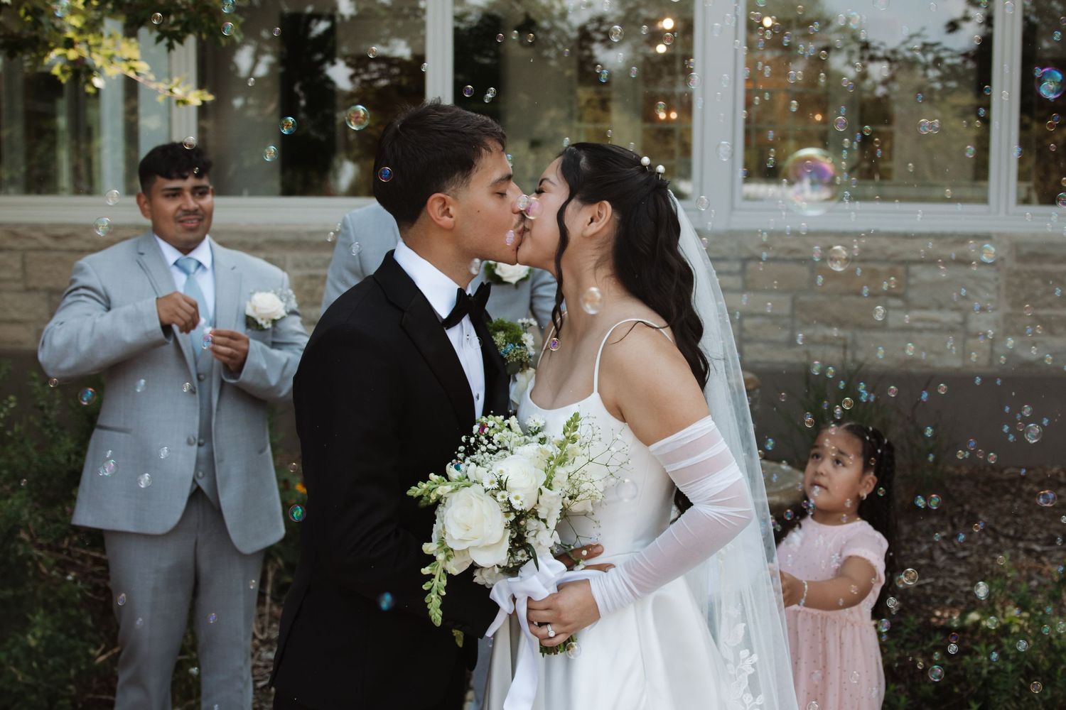 Newlyweds sharing a romantic kiss outdoors surrounded by bubbles with groomsmen and flower girl nearby.