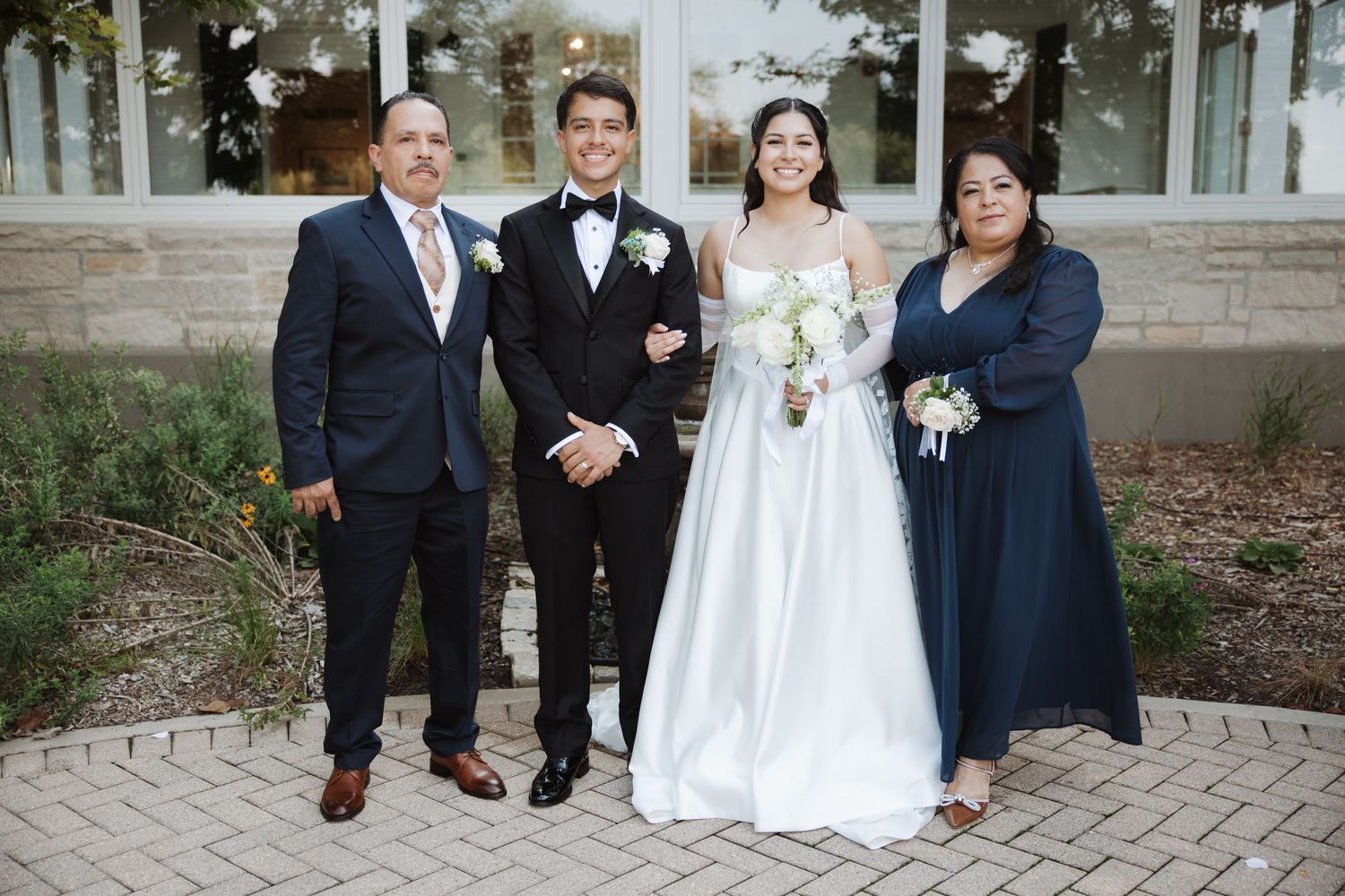 Wedding couple stands with parents in formal attire outside a stone building on their special day.