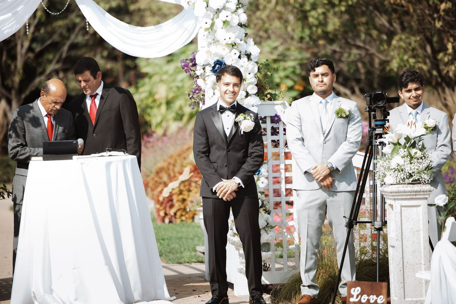 Groom smiling at the altar during an outdoor wedding ceremony with groomsmen and officiant nearby.