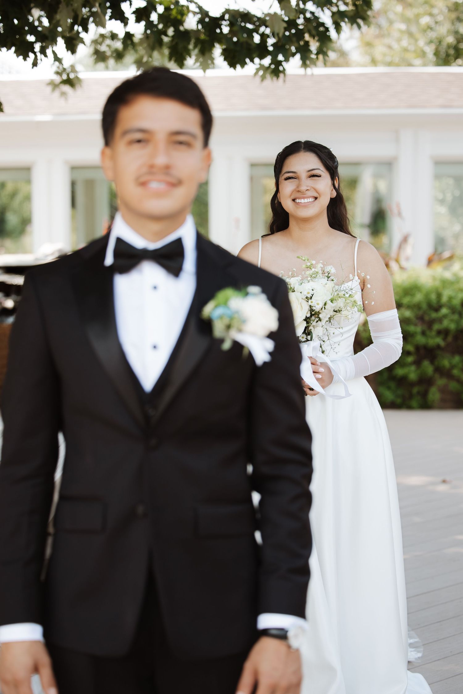 Groom in black tuxedo stands in foreground as smiling bride holds bouquet behind him.