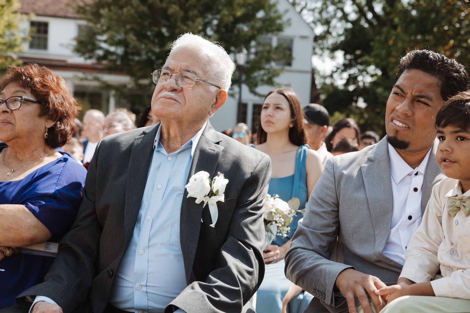 An elderly man in a blazer with a white boutonniere sits beside a younger man at an outdoor wedding.