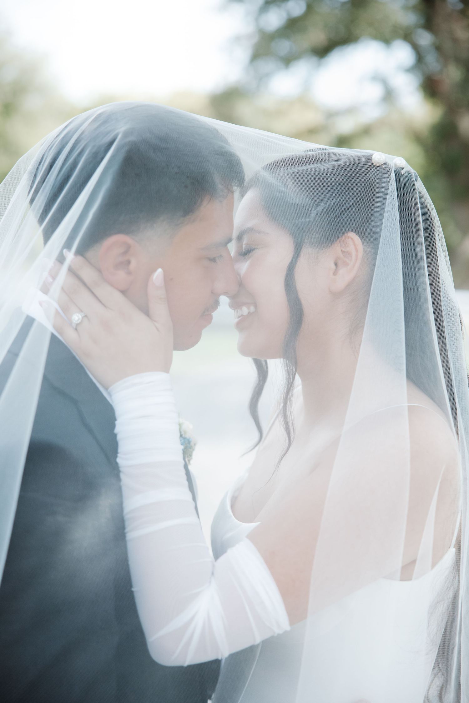 Happy couple sharing a romantic moment under a sheer bridal veil, smiling and holding each other on wedding day.