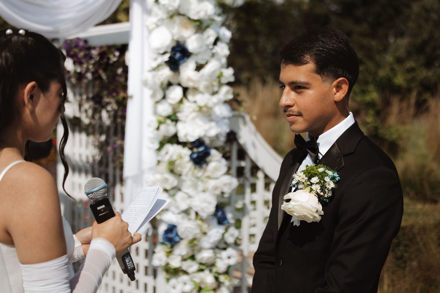 Groom listens attentively as bride reads vows at flower-adorned outdoor wedding ceremony.