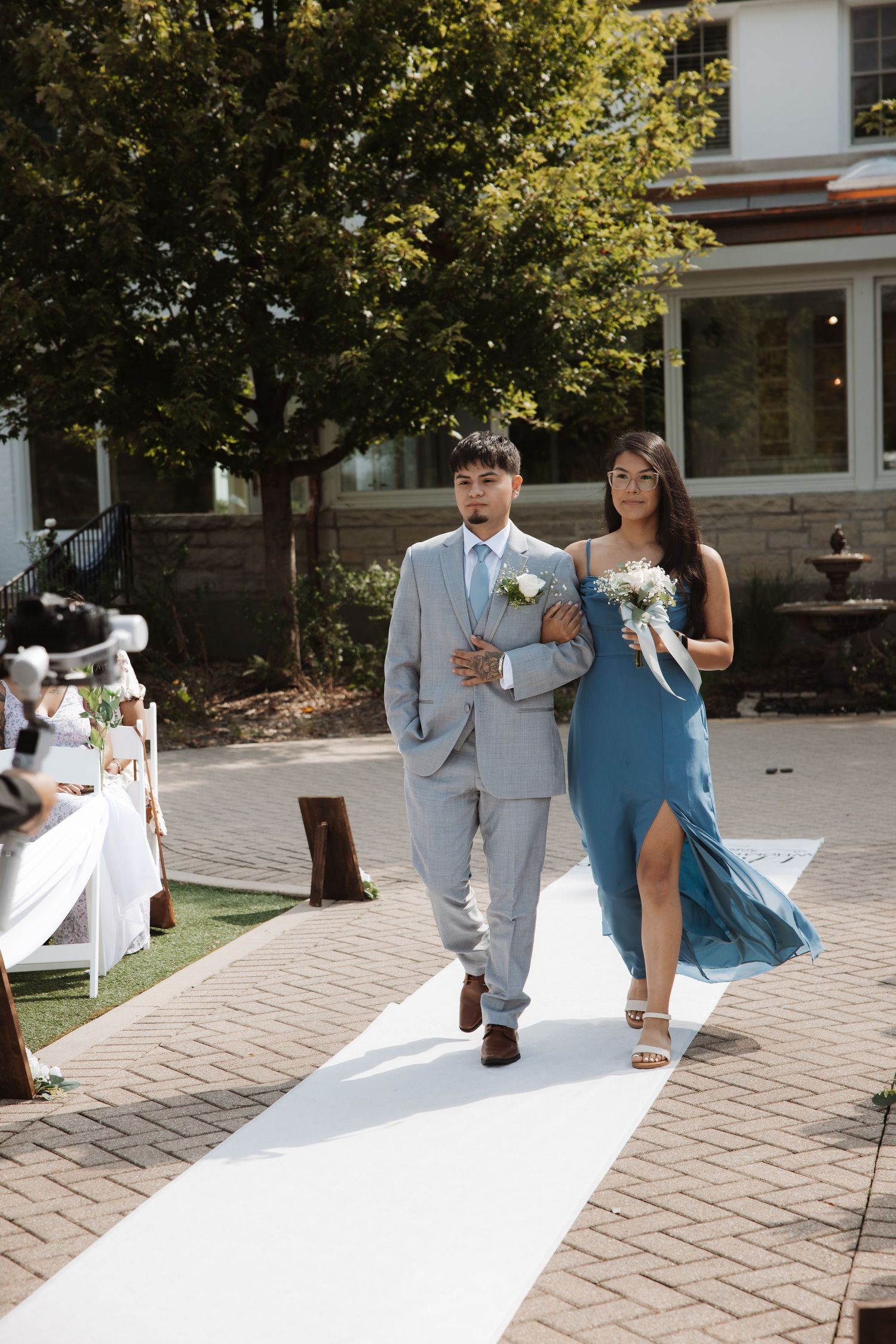 Wedding party members walk down white aisle runner at outdoor ceremony venue.