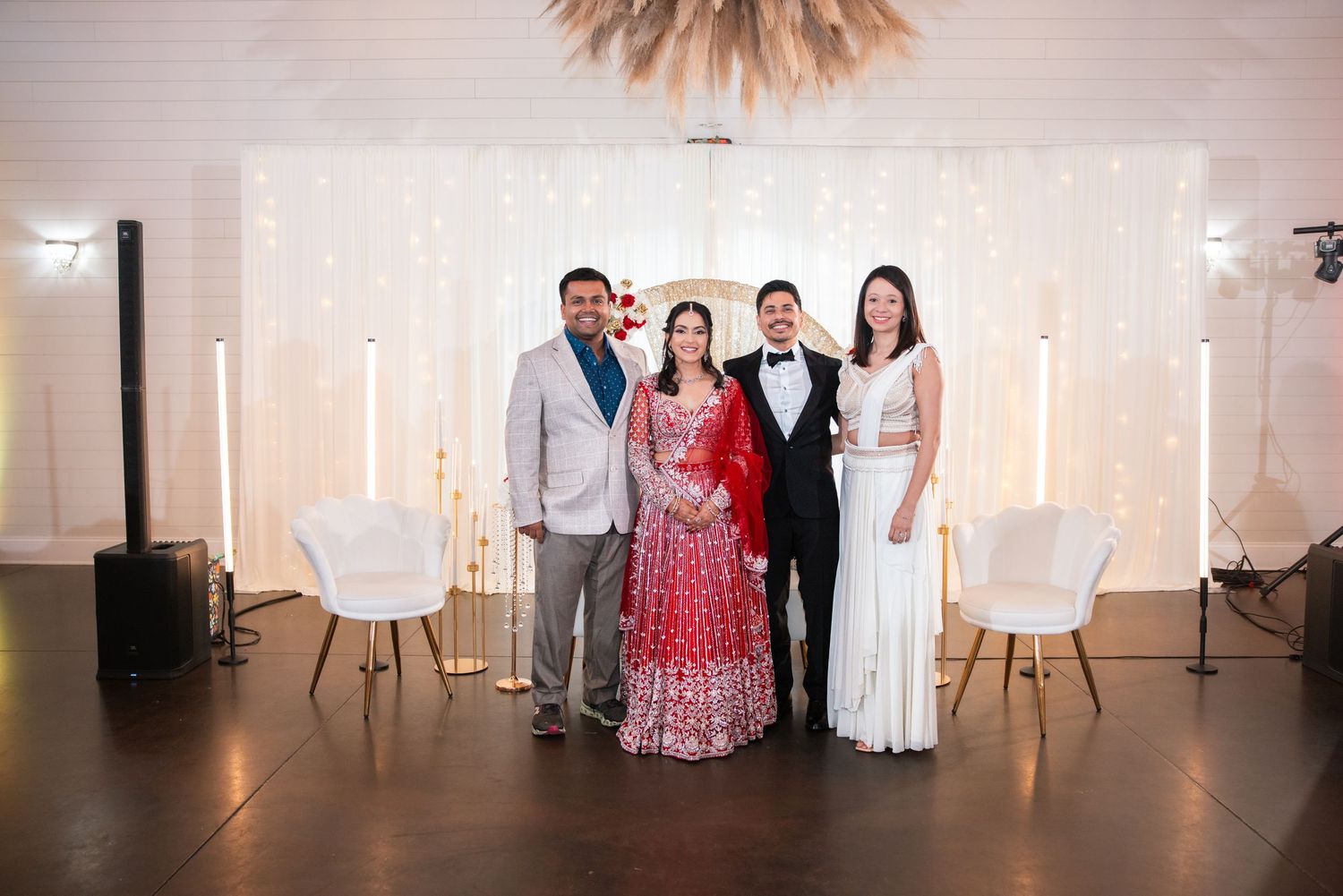 Bride in red lehenga poses with groom in tuxedo and two guests at elegant wedding reception.