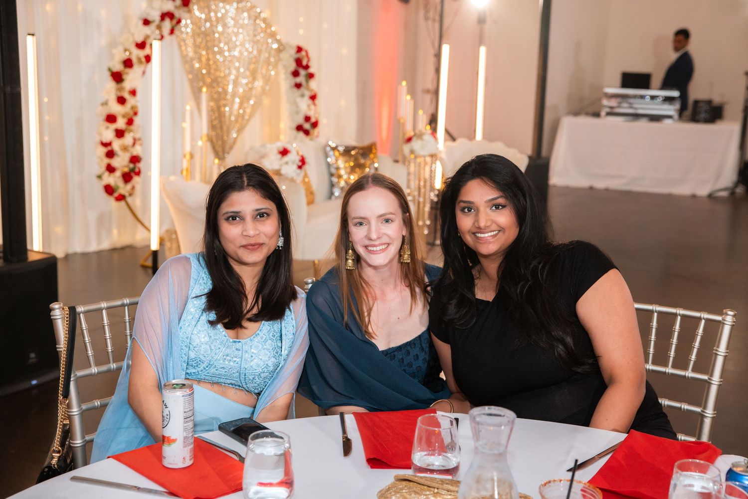 Three women smile together at table decorated for elegant Indian wedding reception.