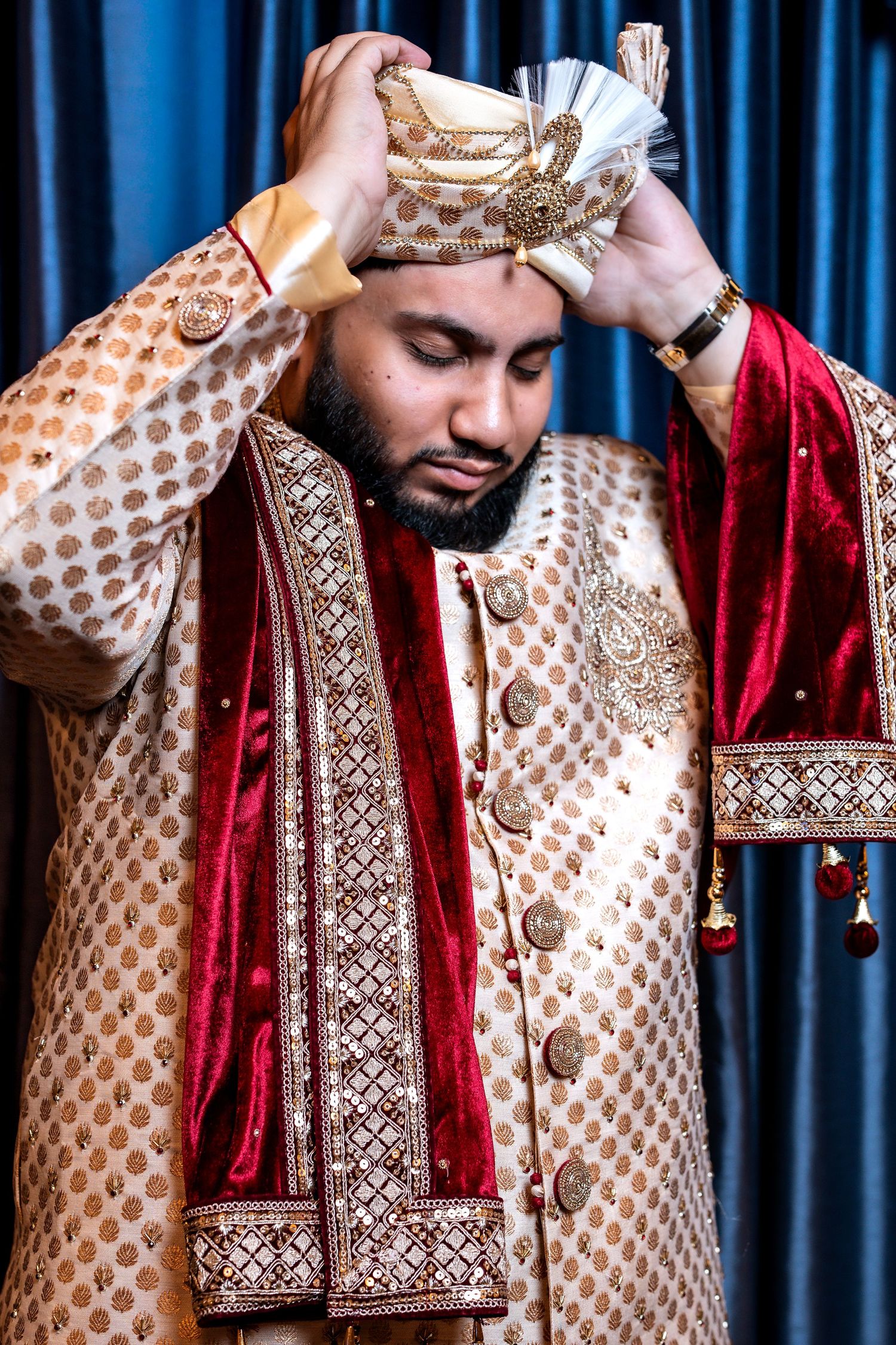 Indian groom in gold sherwani adjusting his decorated turban while standing before blue curtains.