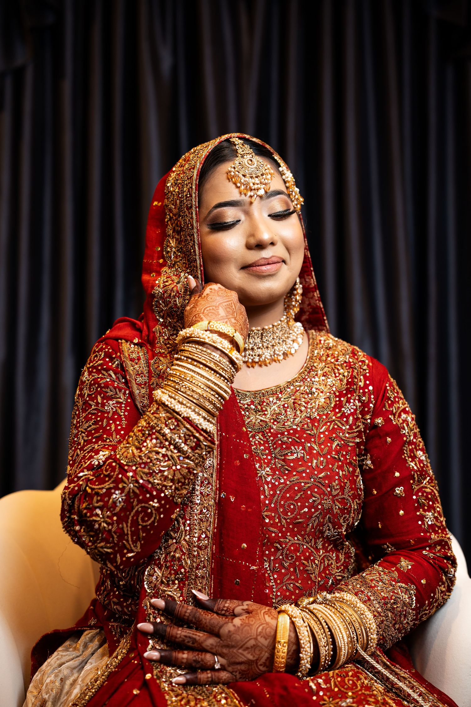 South Asian bride in red bridal lehenga smiles softly with eyes closed wearing gold jewelry.