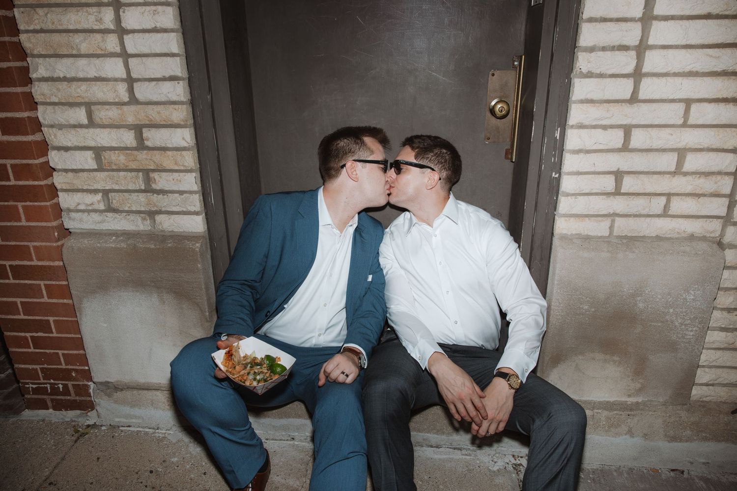 Two grooms in suits sharing a kiss while seated on steps outside a brick building at their wedding.