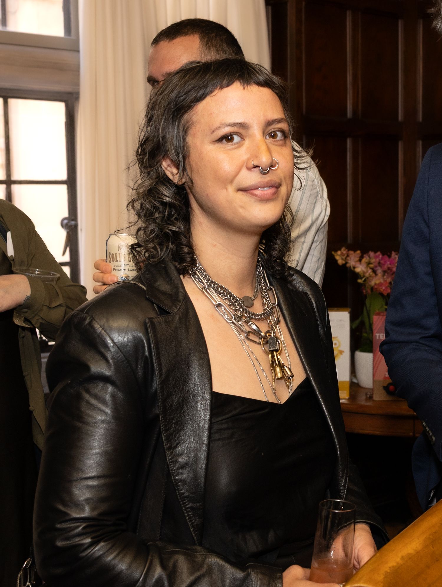 Woman with curly hair wearing a black leather jacket and layered necklaces at a social gathering.