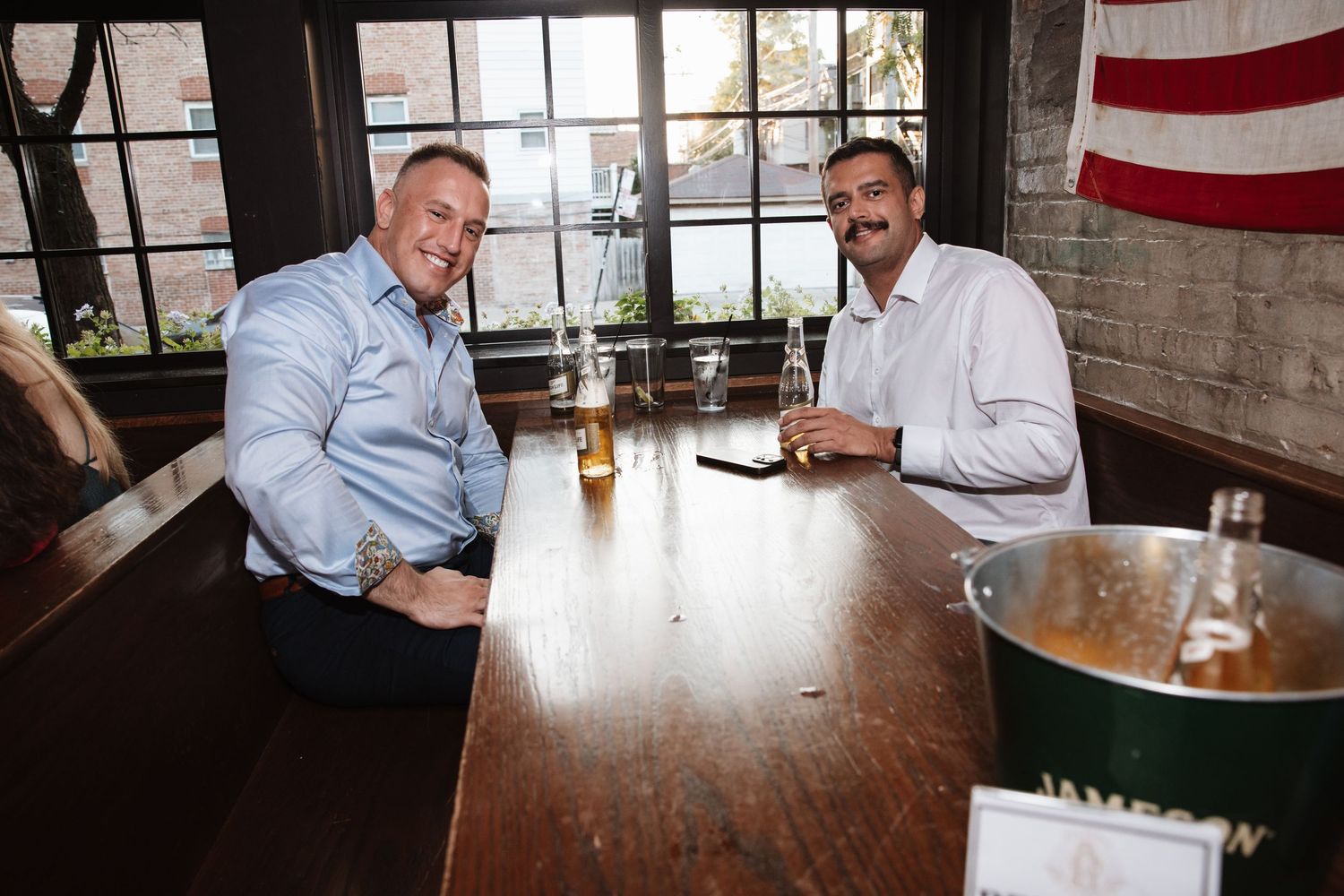 Two men smiling at a wooden bar with drinks and a champagne bucket.