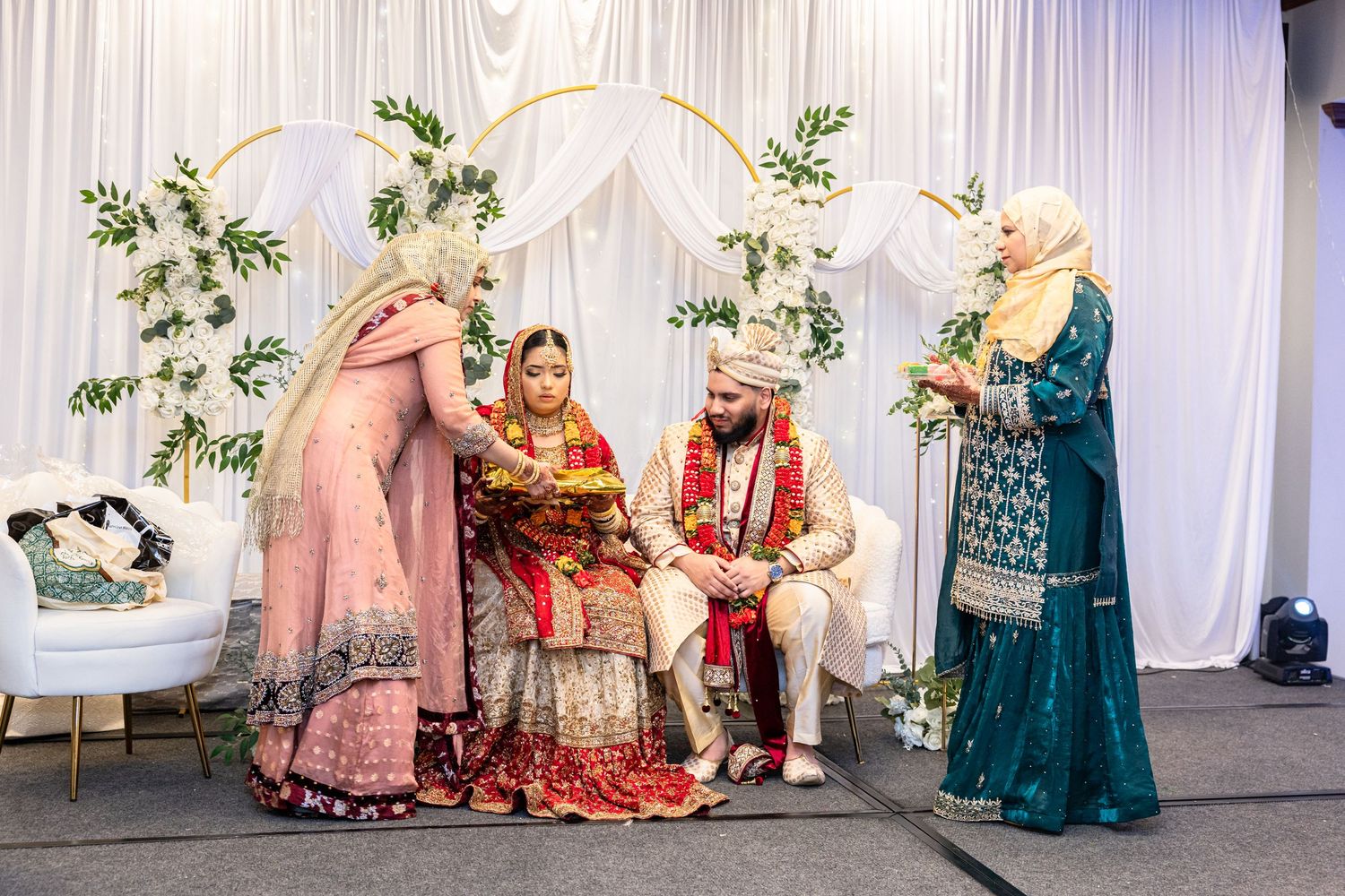 Traditional South Asian wedding ceremony with bride and groom seated under decorative arch.