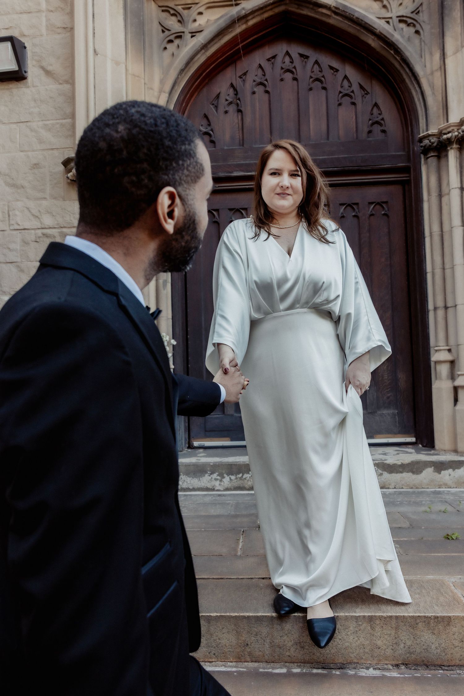 Bride in white gown holds groom's hand on church steps in elegant wedding photo.