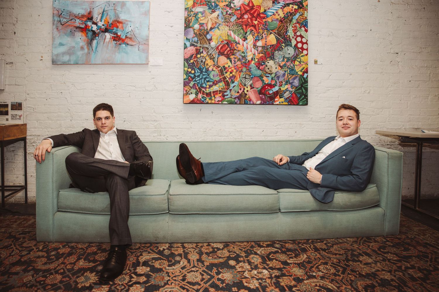 Two men in formal suits relax on a mint green sofa beneath colorful abstract wall art in a loft.