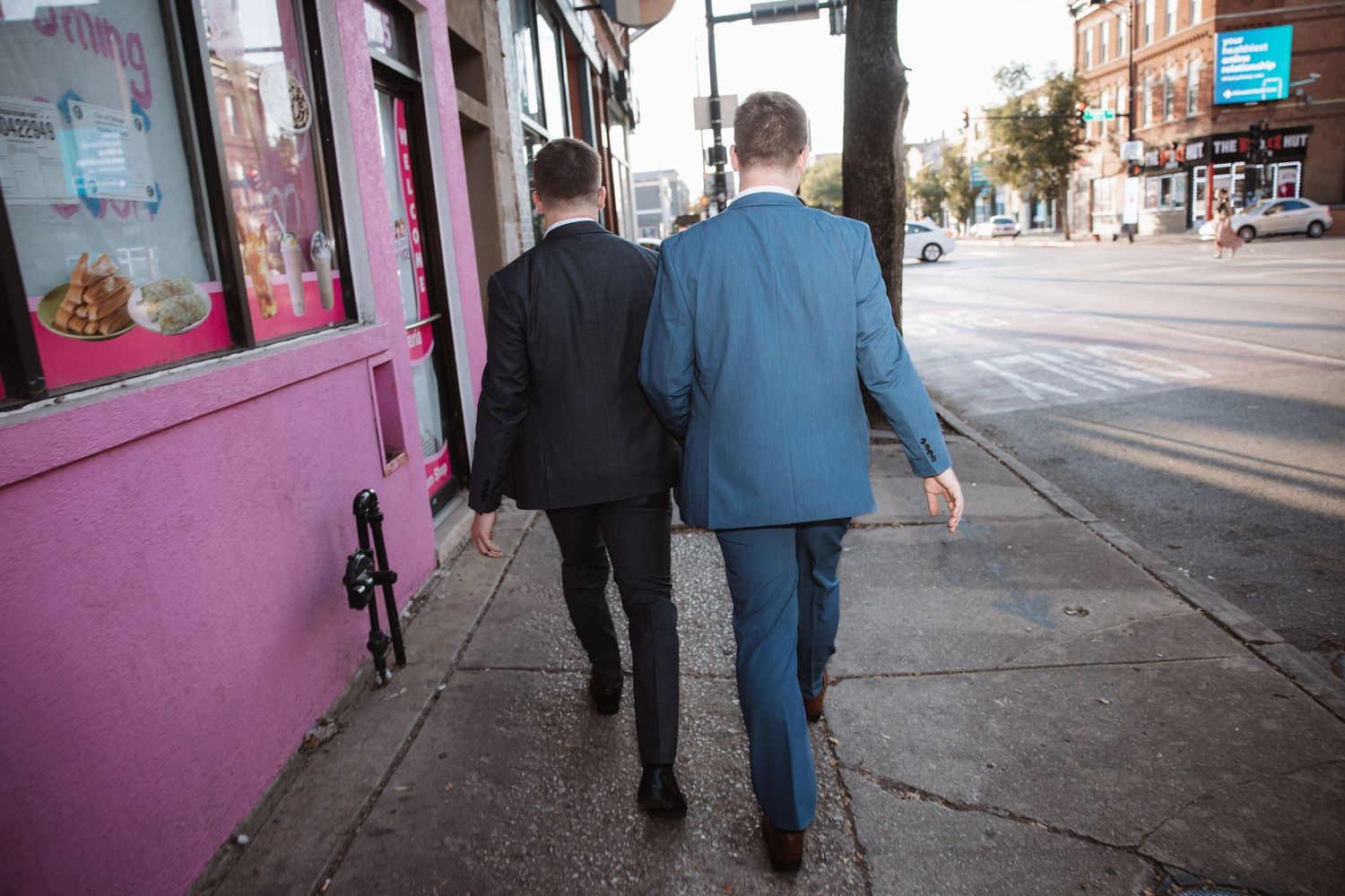 Two grooms in suits walk hand in hand down urban sidewalk on their wedding day.