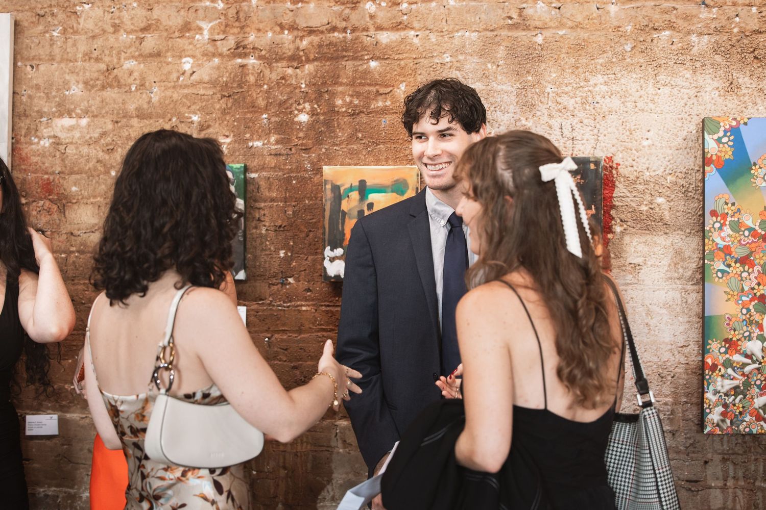 Young man in suit smiling while chatting with two women at an art gallery event.