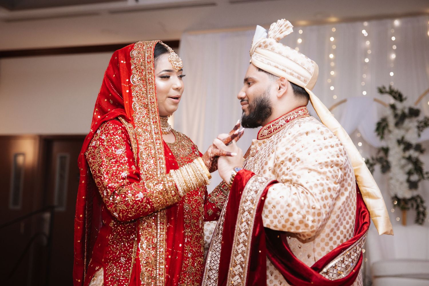 Bride and groom in traditional red and gold Indian wedding attire sharing an intimate moment during ceremony.