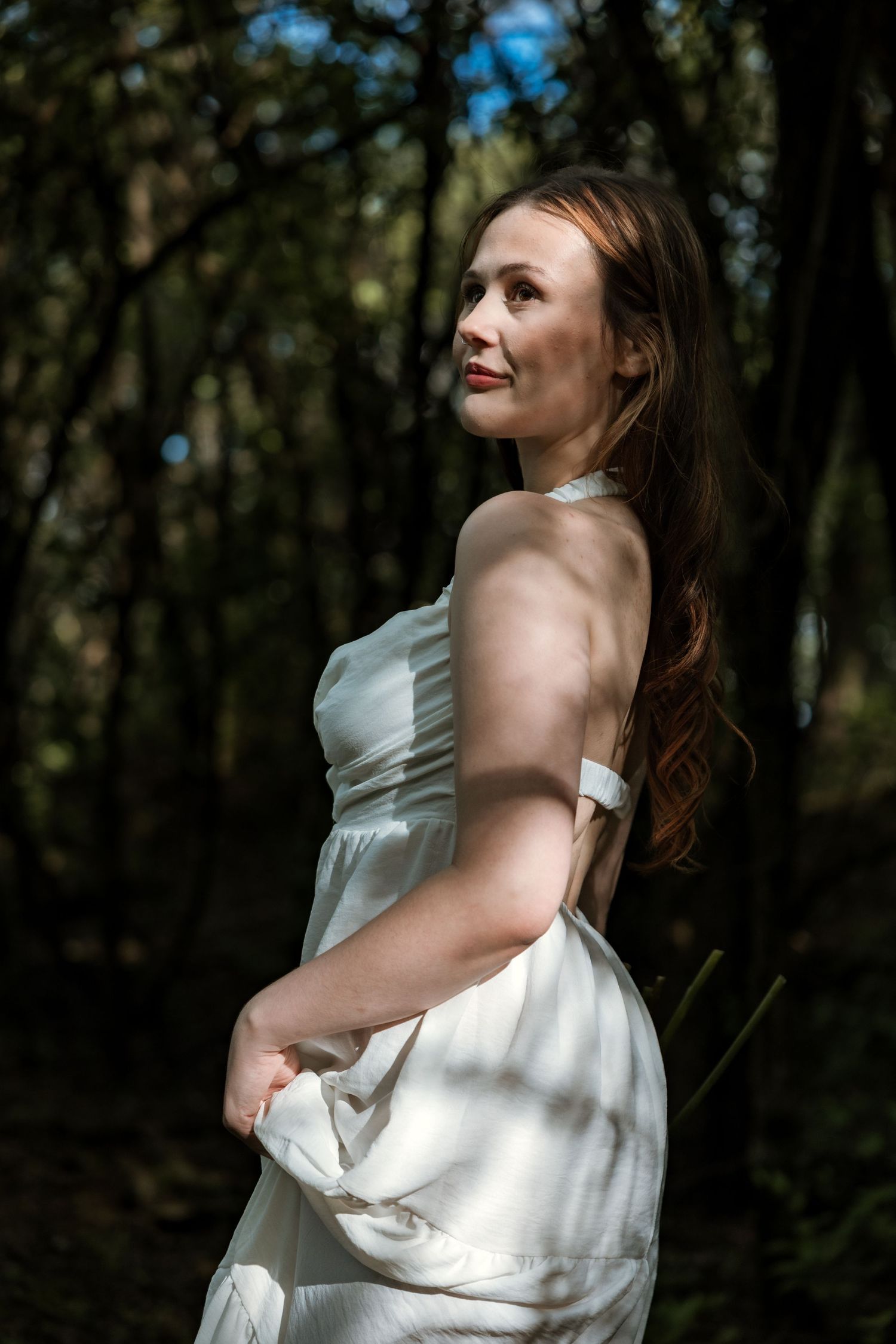 Woman in white satin halter dress smiles softly in dappled sunlight among dark trees.