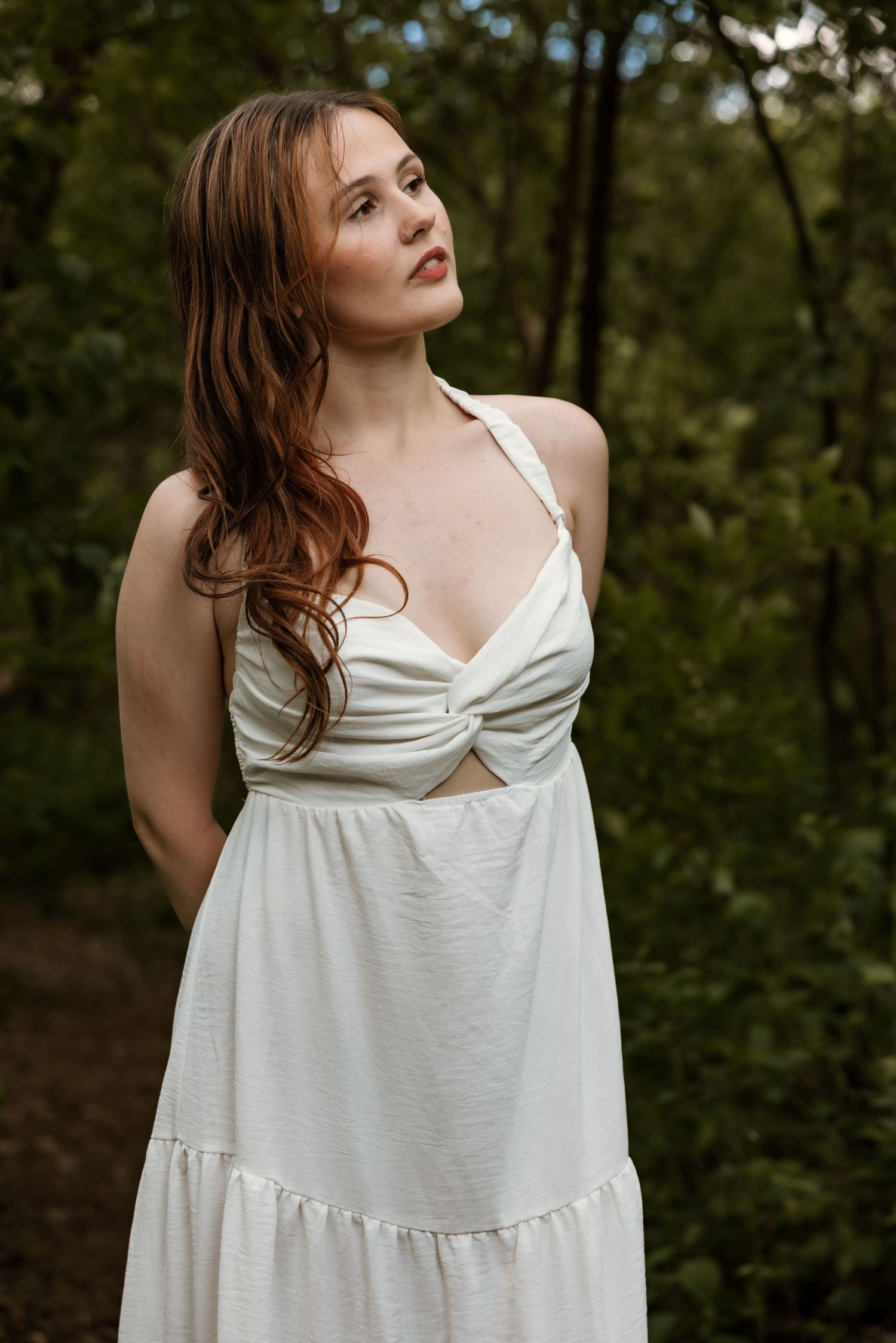 Beautiful redhead woman in white sundress gazes upward among green trees outdoors.