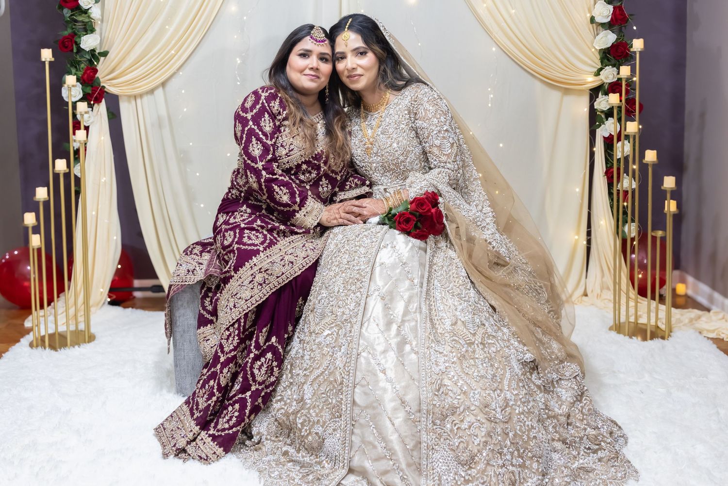 Bride in gold embroidered lehenga poses with woman in purple velvet outfit at elegant wedding backdrop.
