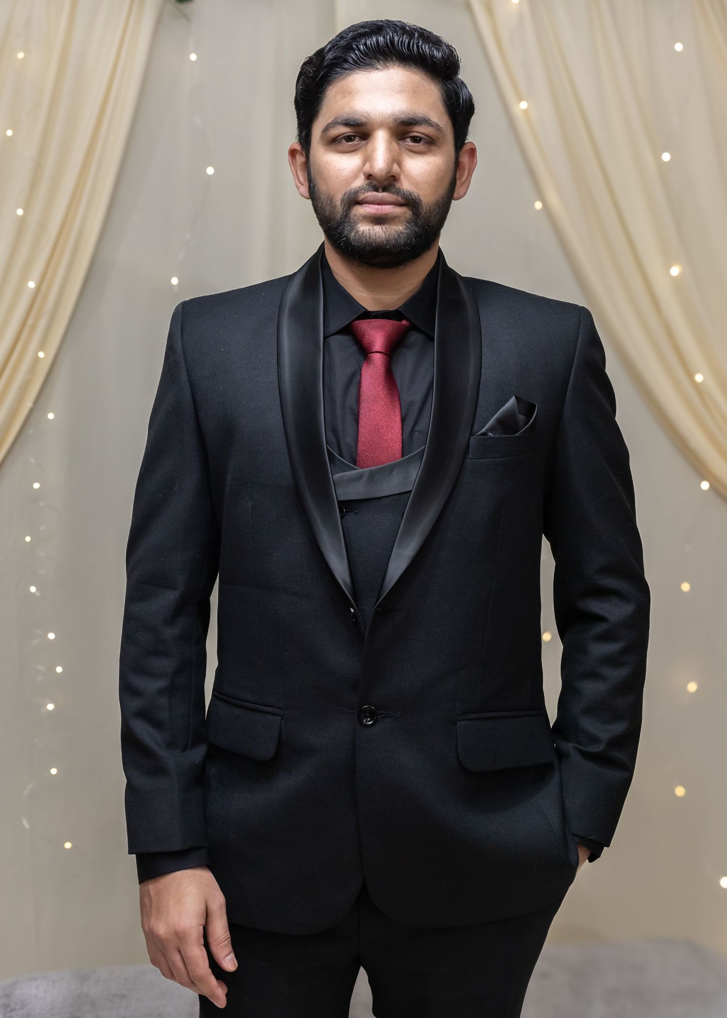 A man in a black tuxedo with a red tie poses confidently at a wedding venue with draped curtains.