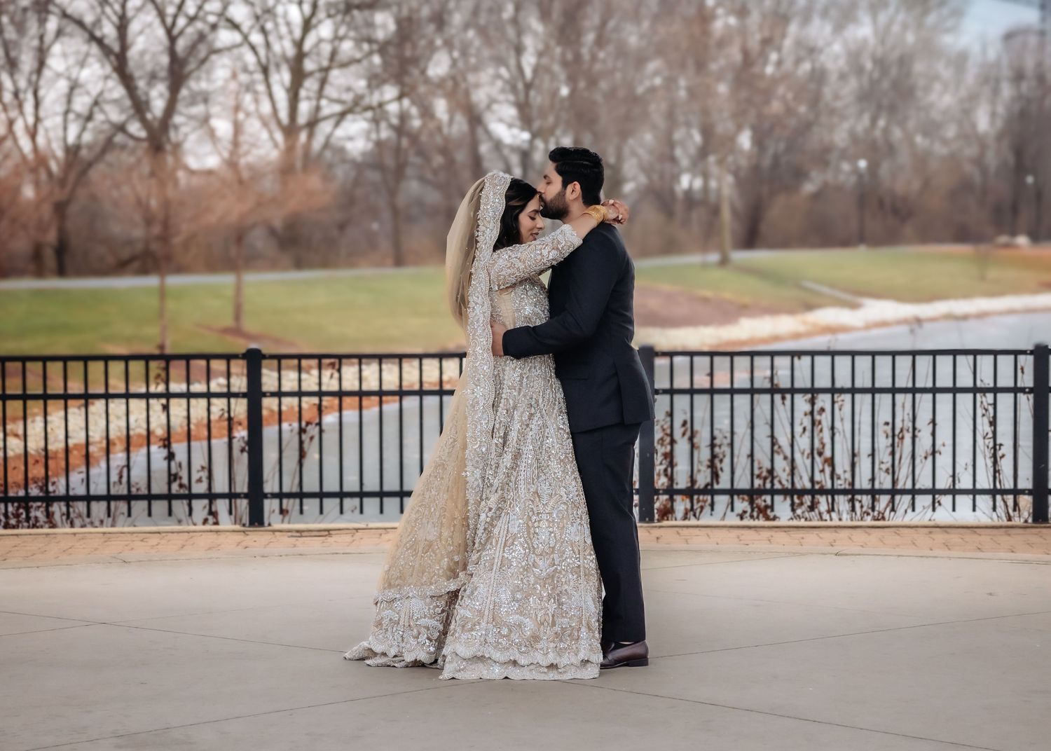 South Asian bride and groom embracing in gold bridal lehenga and navy suit outdoors.