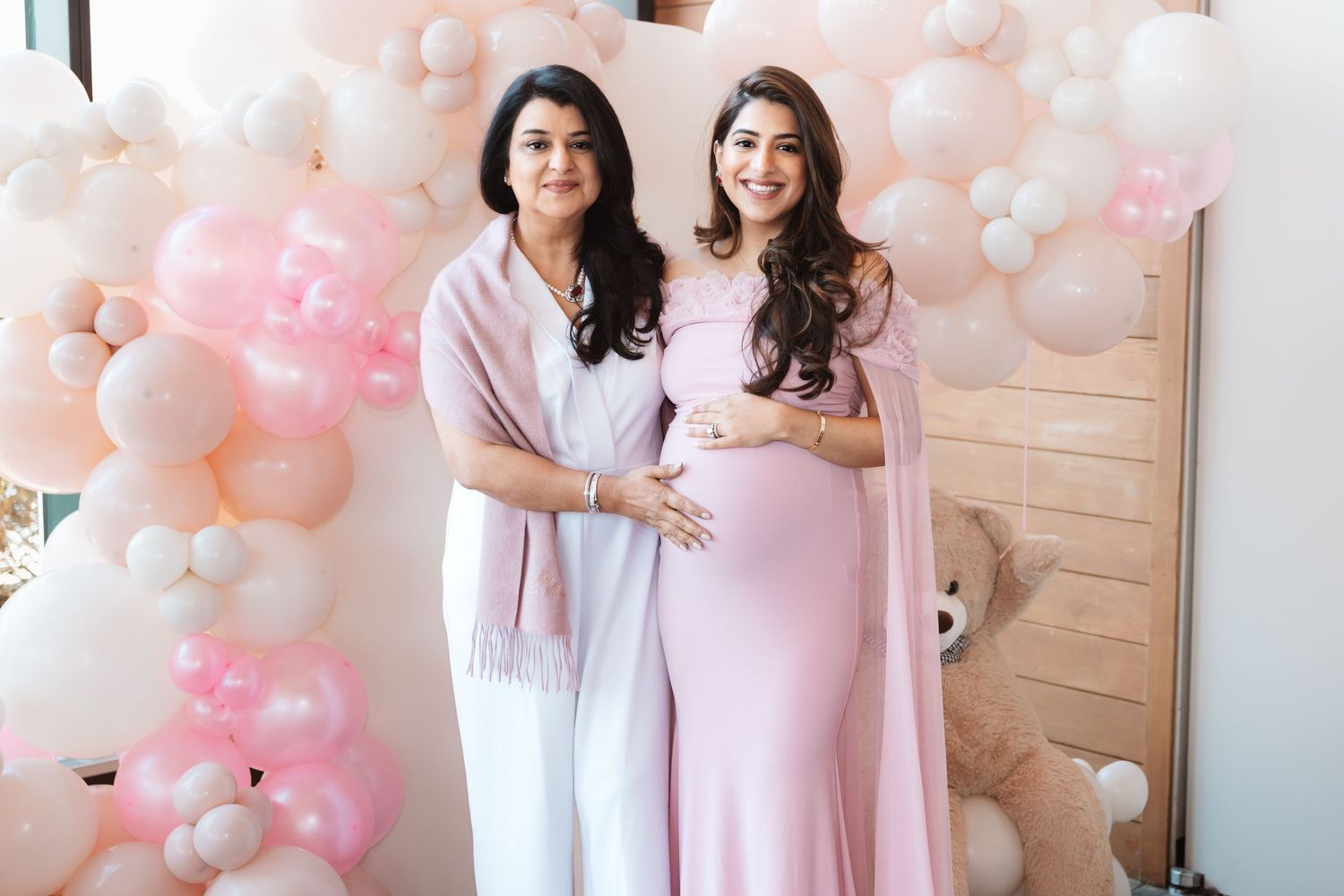 Pregnant South Asian woman in lavender maternity gown poses with mother at pink balloon baby shower.