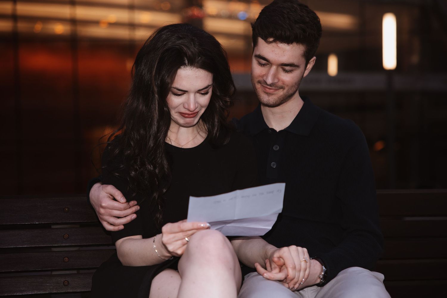Young couple sitting on a bench together reading a letter in a warmly lit urban setting.