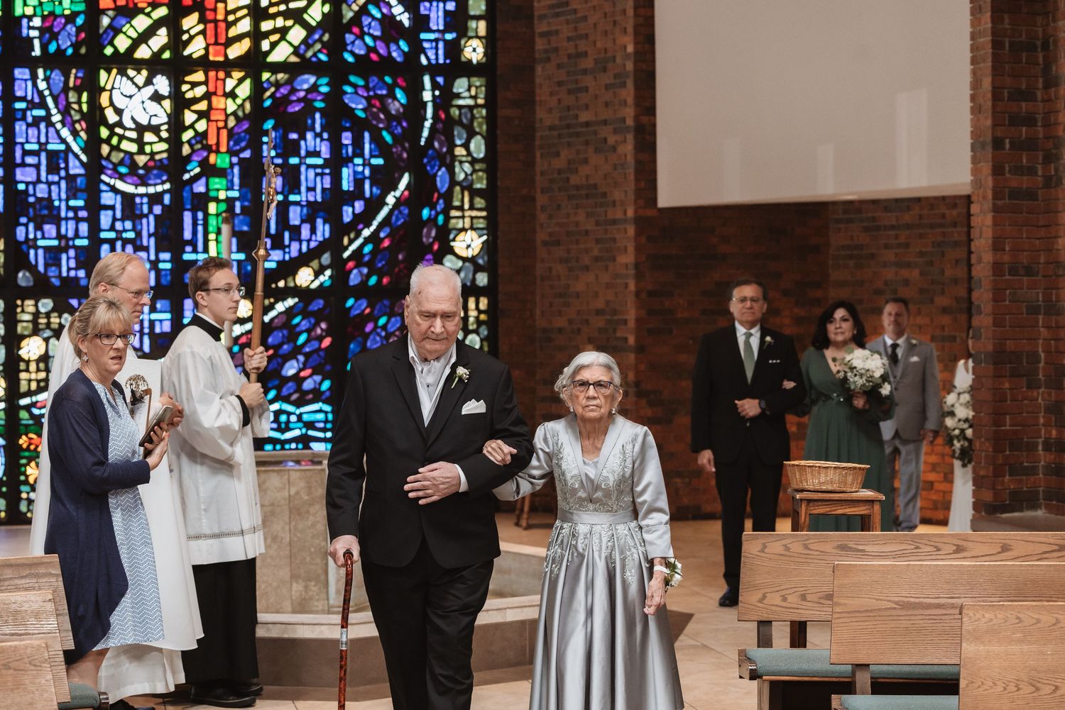 Elderly couple walking down church aisle during wedding ceremony with stained glass windows in background.