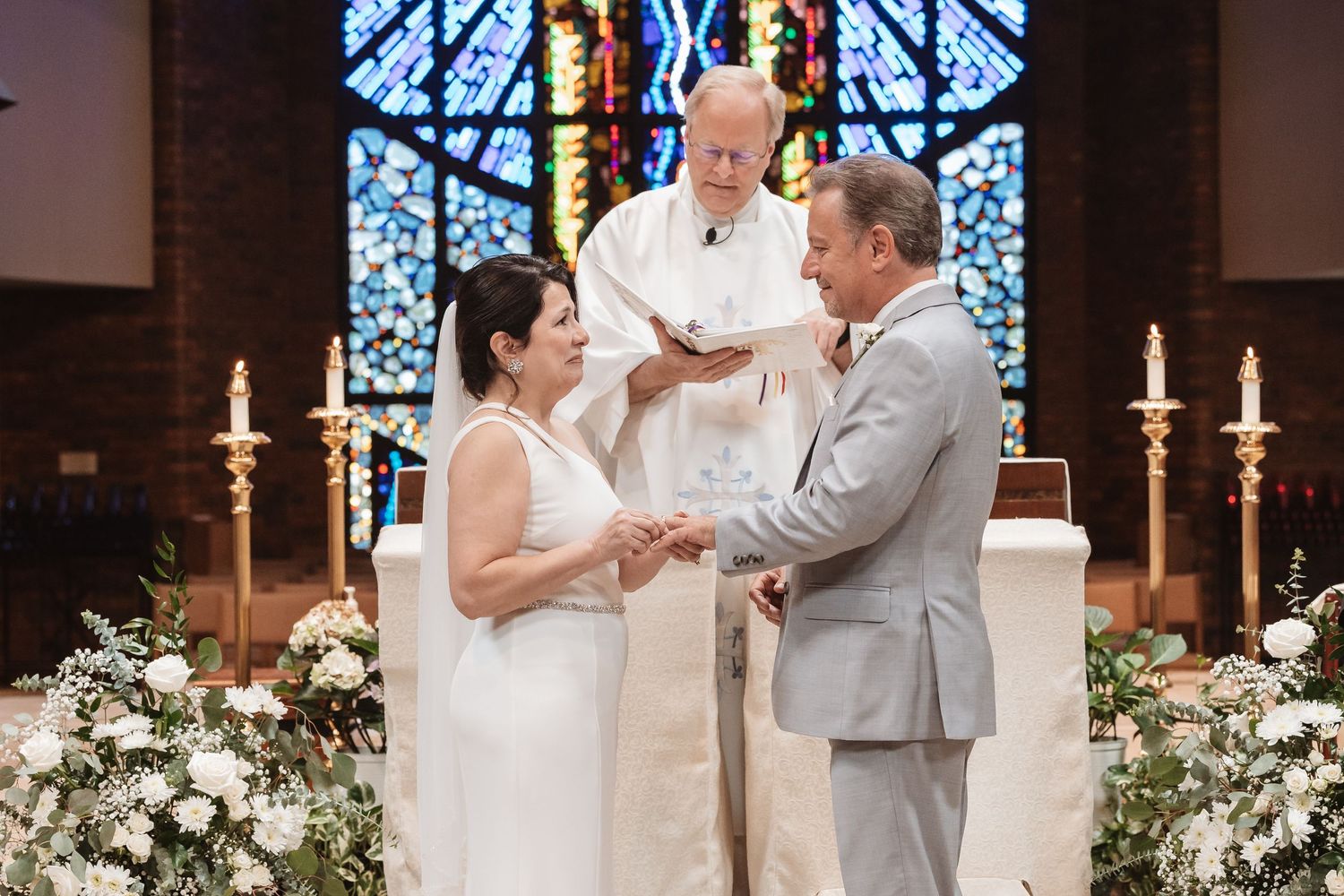 Couple exchanging rings at church altar with officiant during elegant wedding ceremony with white floral arrangements.