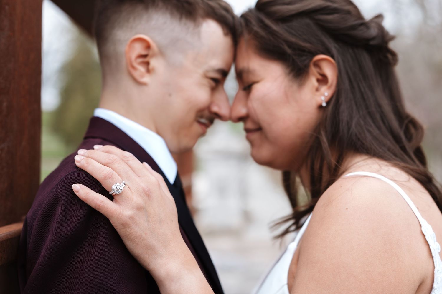 Two brides touching foreheads and smiling, one wearing a burgundy suit and diamond ring on wedding day.