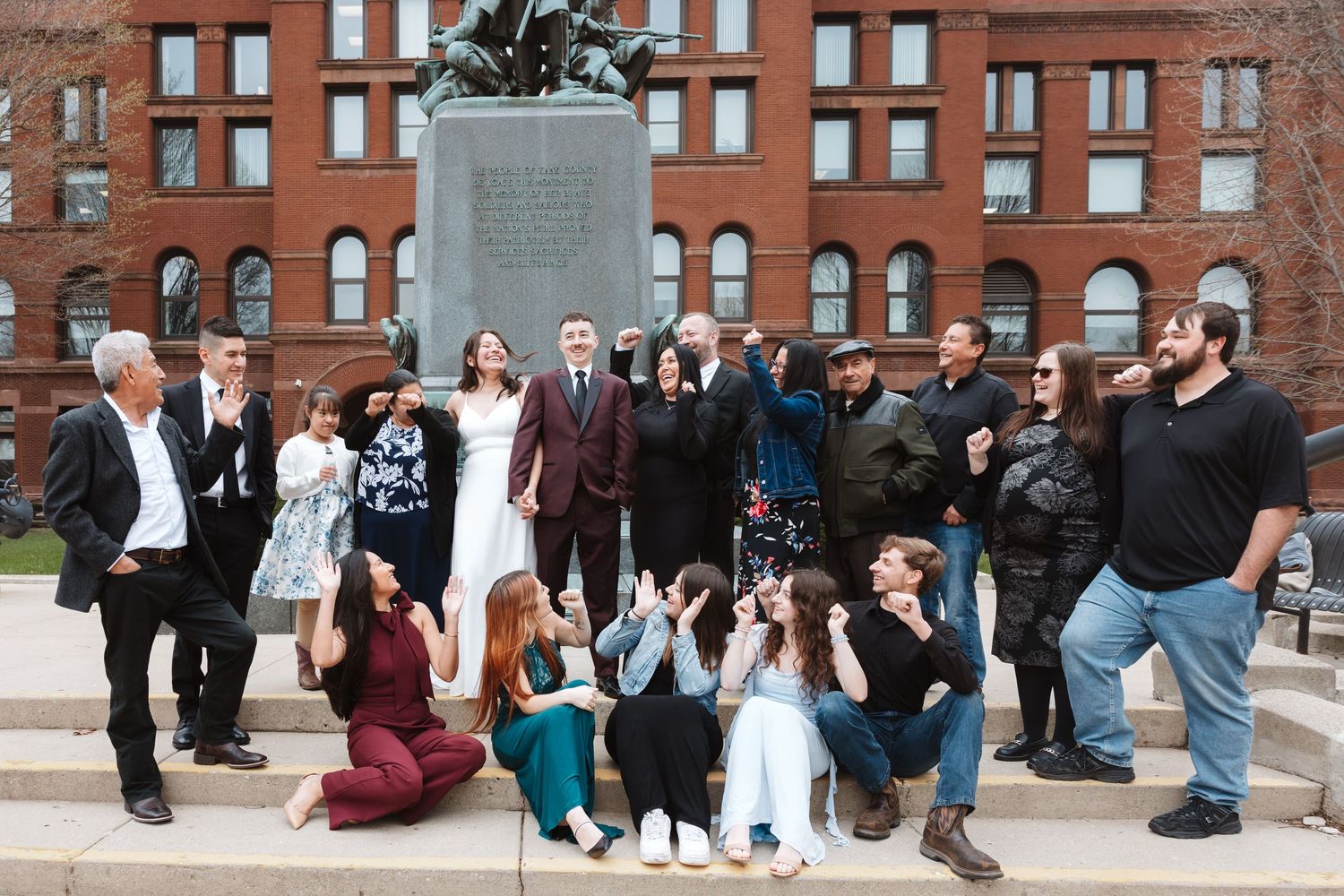 Large wedding party celebrating outdoors near a bronze statue in front of a red brick building.