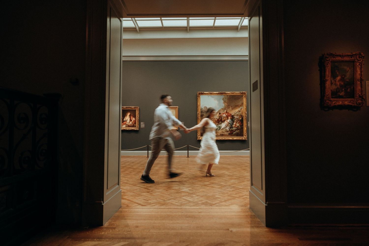 Couple dancing joyfully through elegant museum gallery doorway with classic paintings on walls and warm parquet floors.